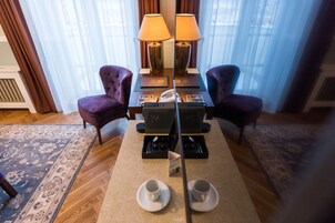 Quarto de hotel com piso de madeira, duas poltronas roxas, uma mesa com duas luminárias, um telefone e duas xícaras com pires.