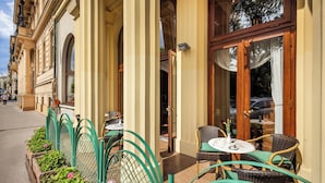 Un café al aire libre con sillas y mesas de mimbre, un macizo de flores rosas y un edificio con grandes columnas y ventanas arqueadas.