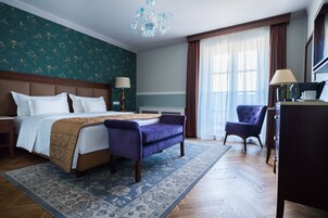Quarto de hotel com uma cama grande, duas mesas de cabeceira, uma cadeira, uma escrivaninha e um lustre.