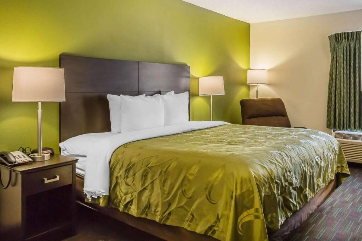 Habitación de hotel con una cama, dos lámparas de noche, una silla, una mesita de noche con un teléfono y una ventana con cortinas.