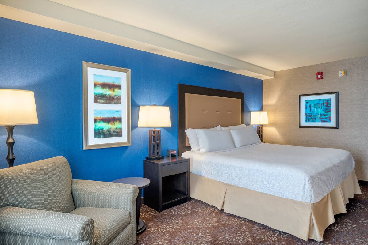 A hotel room with a bed, two bedside lamps, a chair, and two framed pictures on the wall.