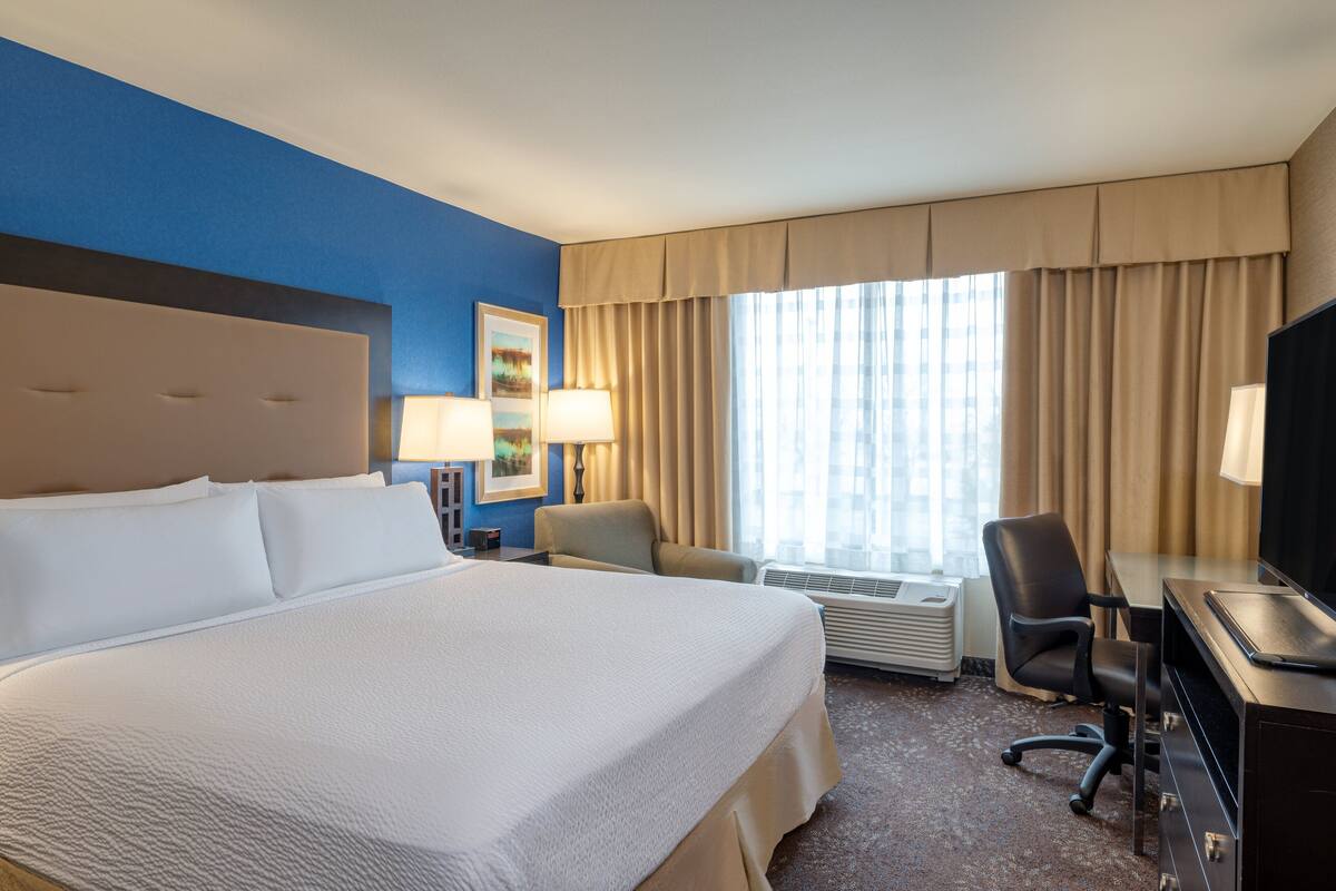 A hotel room with a large bed, a desk with a computer, a chair, a lamp, and a window with curtains.