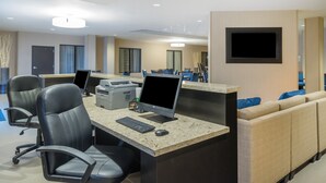 A modern hotel lobby with multiple workstations, a seating area, and a flat-screen TV mounted on the wall. - Holiday Inn Terre Haute by IHG (Terre Haute)