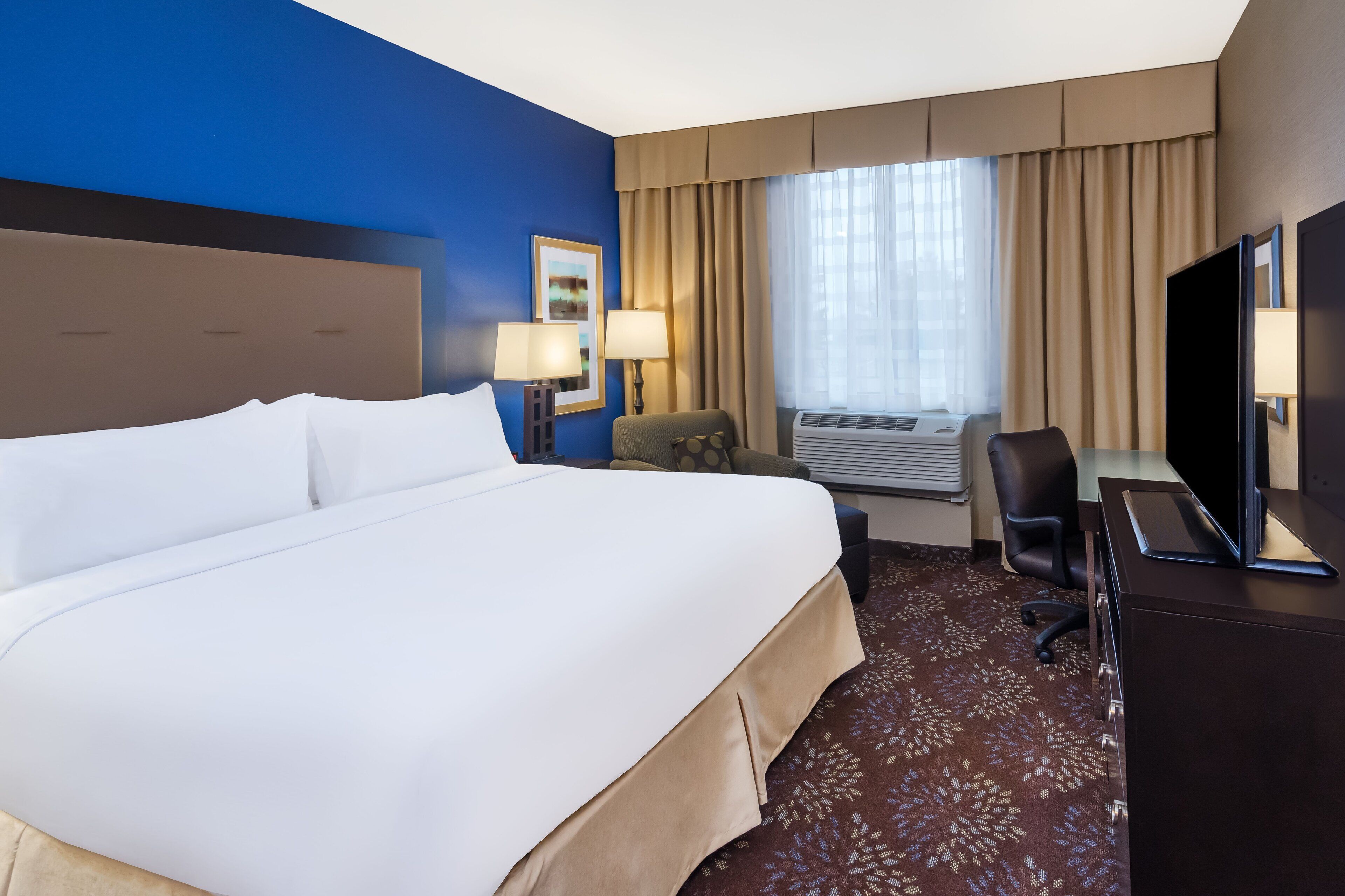 A hotel room with a large bed, two bedside lamps, a desk with a TV, a chair, and an air conditioning unit.