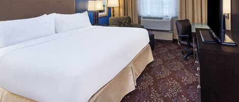 A hotel room with a large bed, two bedside lamps, a desk with a TV, a chair, and an air conditioning unit.