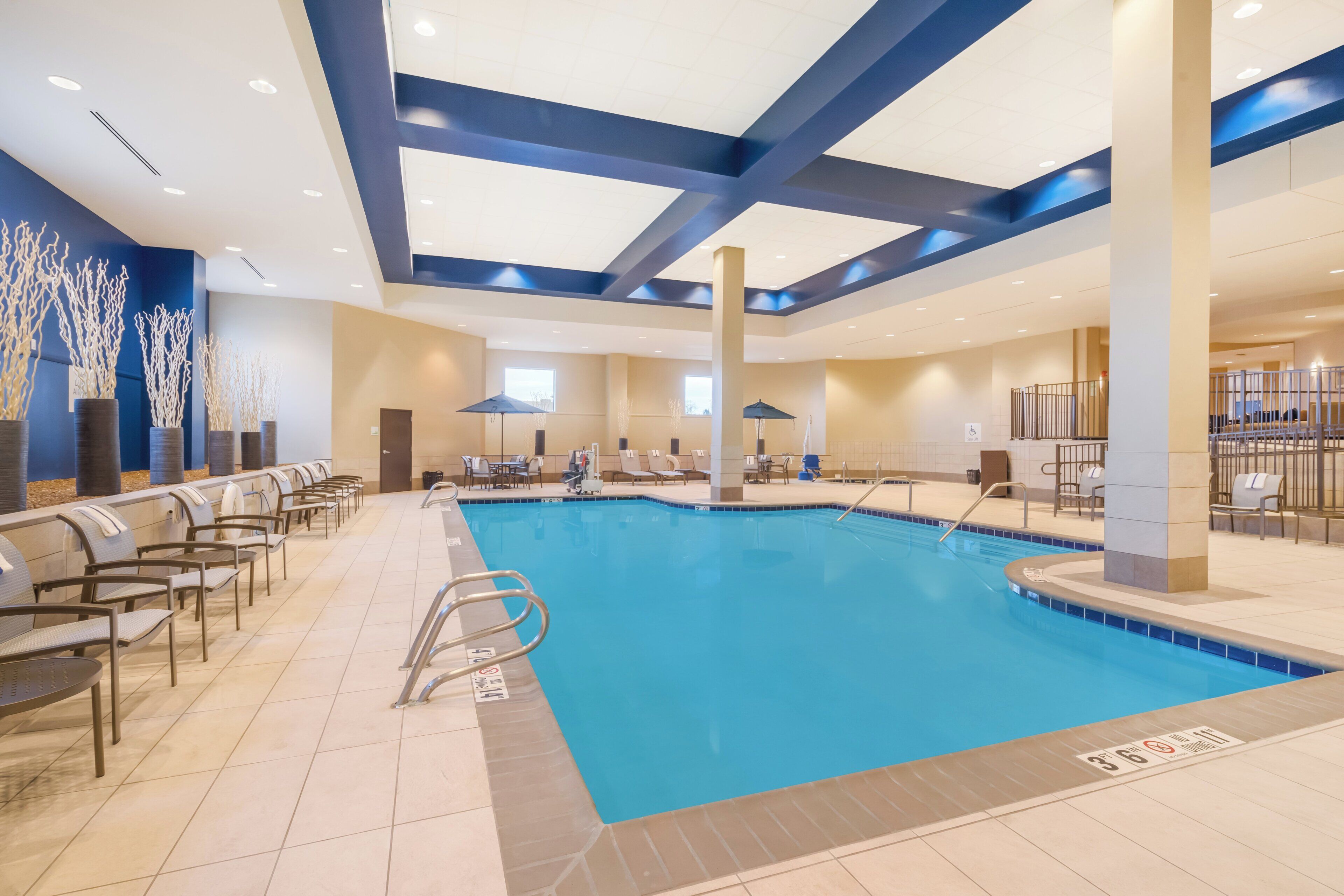 Una piscina con sillones de descanso y sombrillas, rodeada de paredes color beige y detalles en azul.