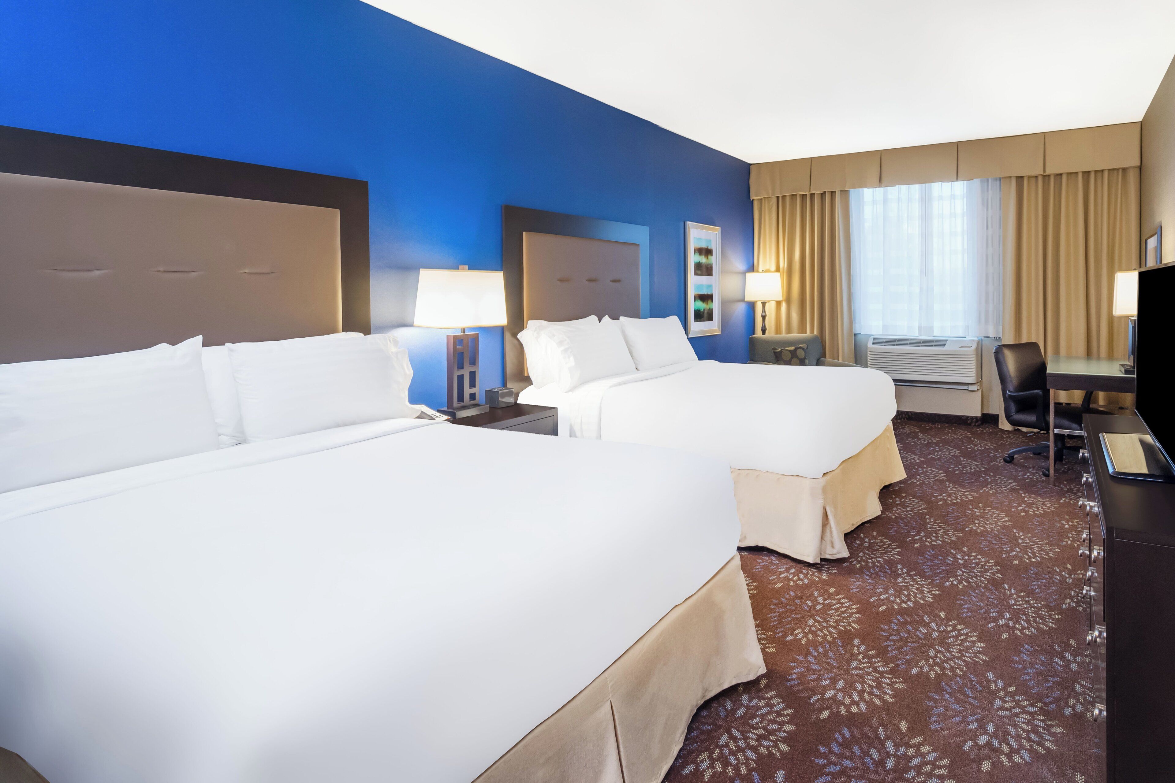 A hotel room with two beds, a desk, a chair, and a large window with curtains.