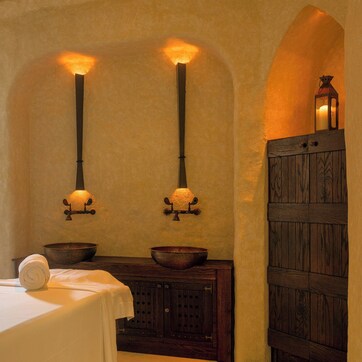 A spa room with a massage table, two wall-mounted candles, a wooden cabinet, and a small window with a view.