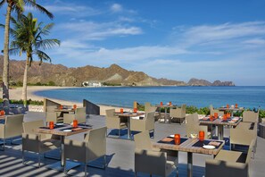 Un área de comedor al aire libre con mesas y sillas, con vista a la playa y montañas.
