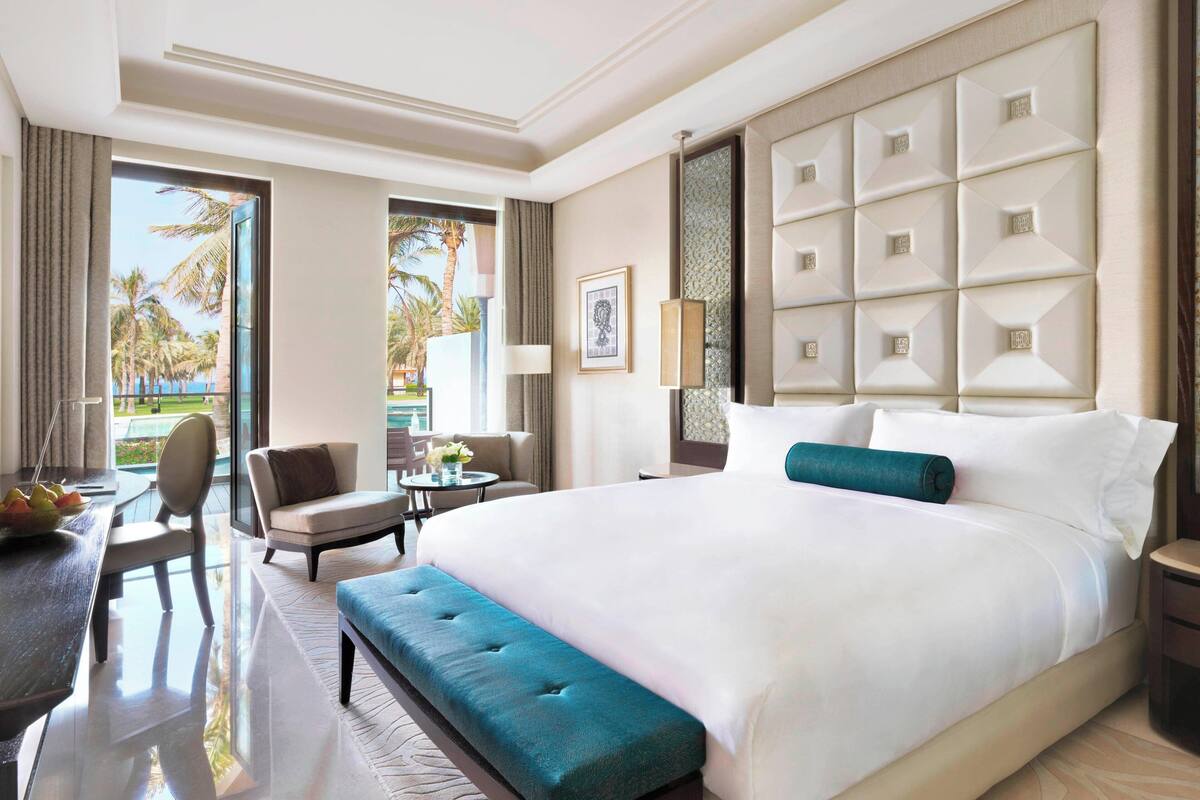 A hotel room with a large bed, a sitting area with a table and chairs, and a view of palm trees through the window.