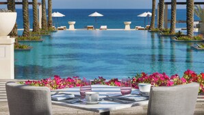 A dining area with a view of a pool and the sea, surrounded by palm trees and flowers.