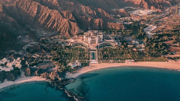 An aerial view of a coastal resort with a central building surrounded by mountains, a beach, and a marina.