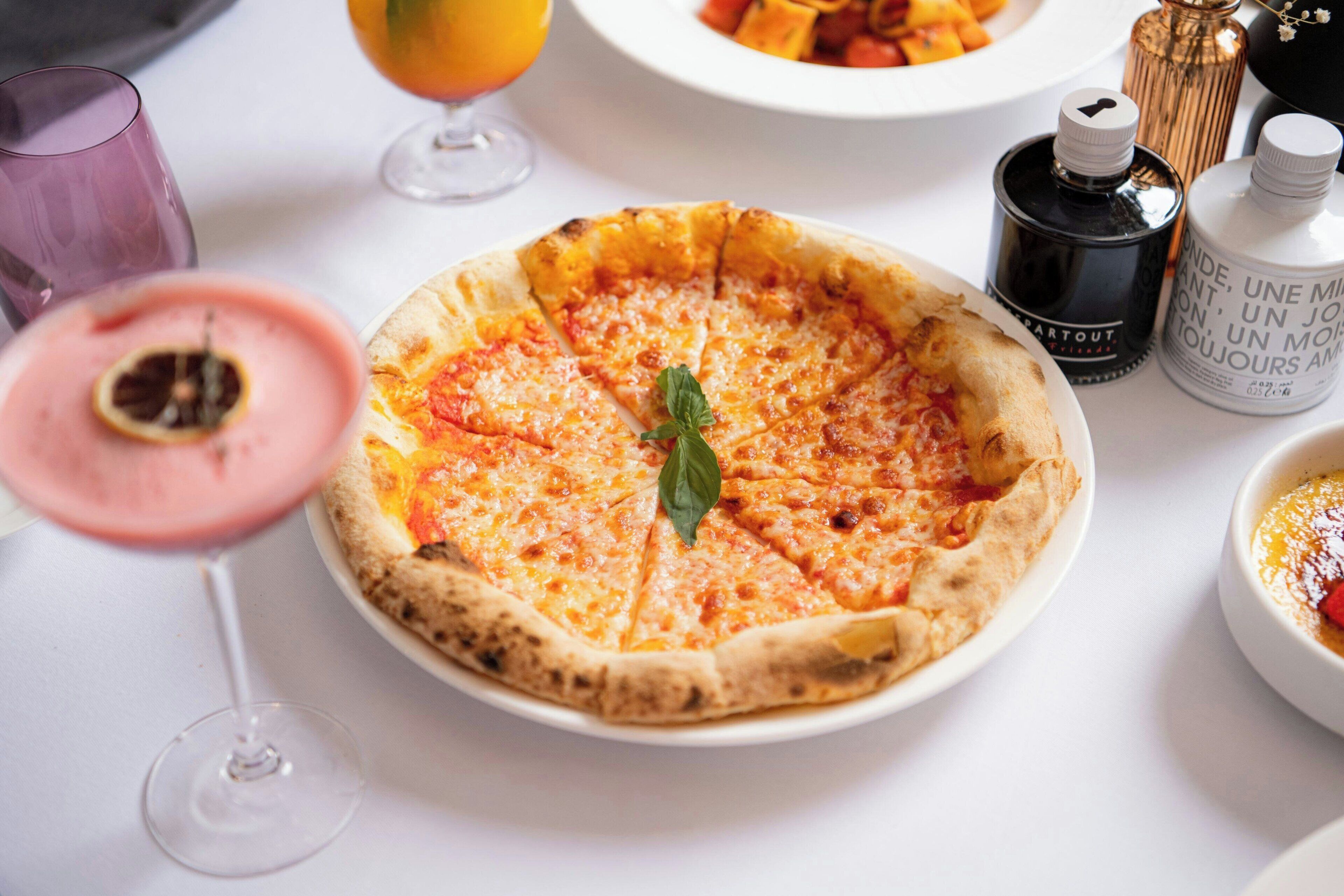 Une pizza sur une assiette blanche, avec un verre de boisson rose et un verre de jus d’orange en arrière-plan.
