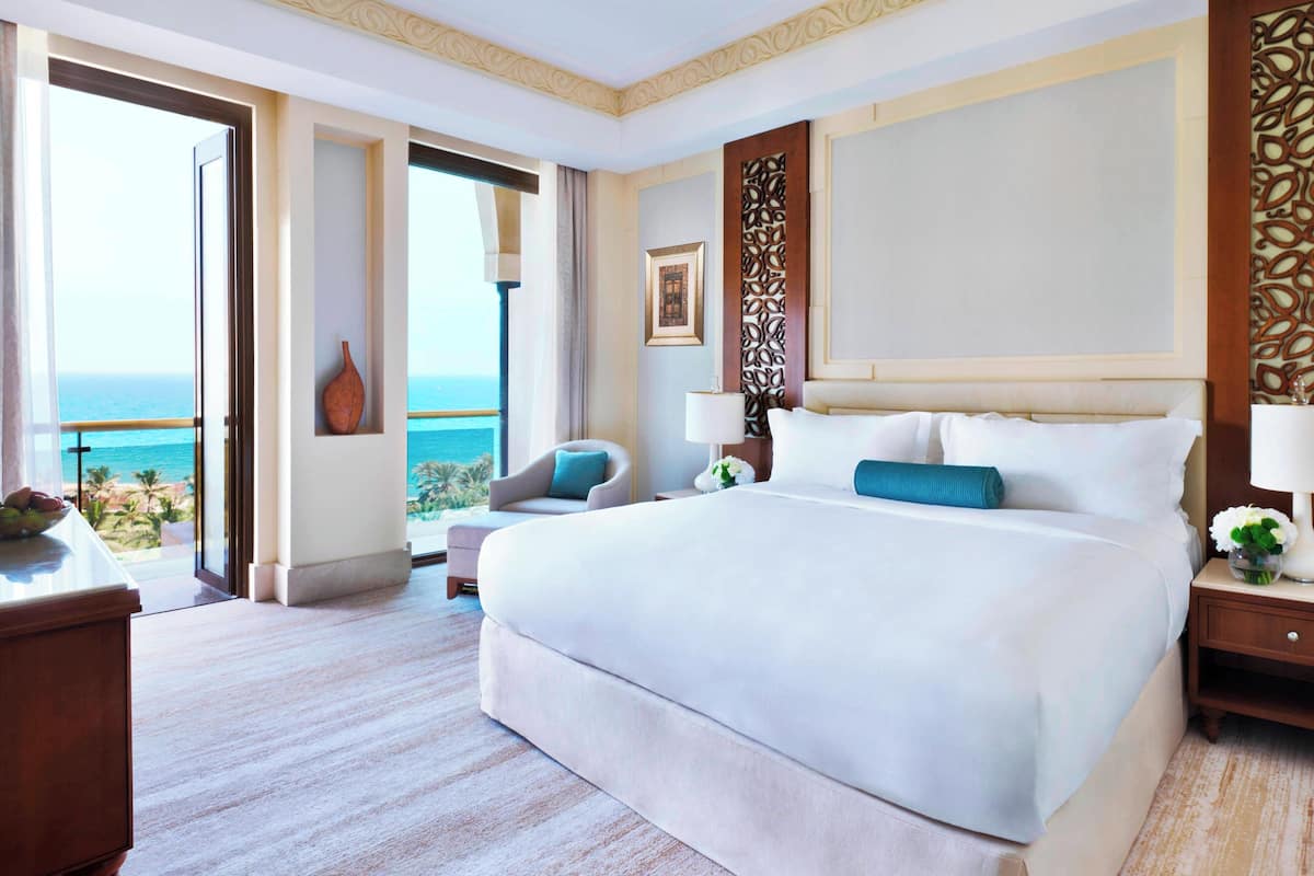 A spacious hotel room with a large bed, a sitting area with a chair and a table, a nightstand, and a view of the ocean.