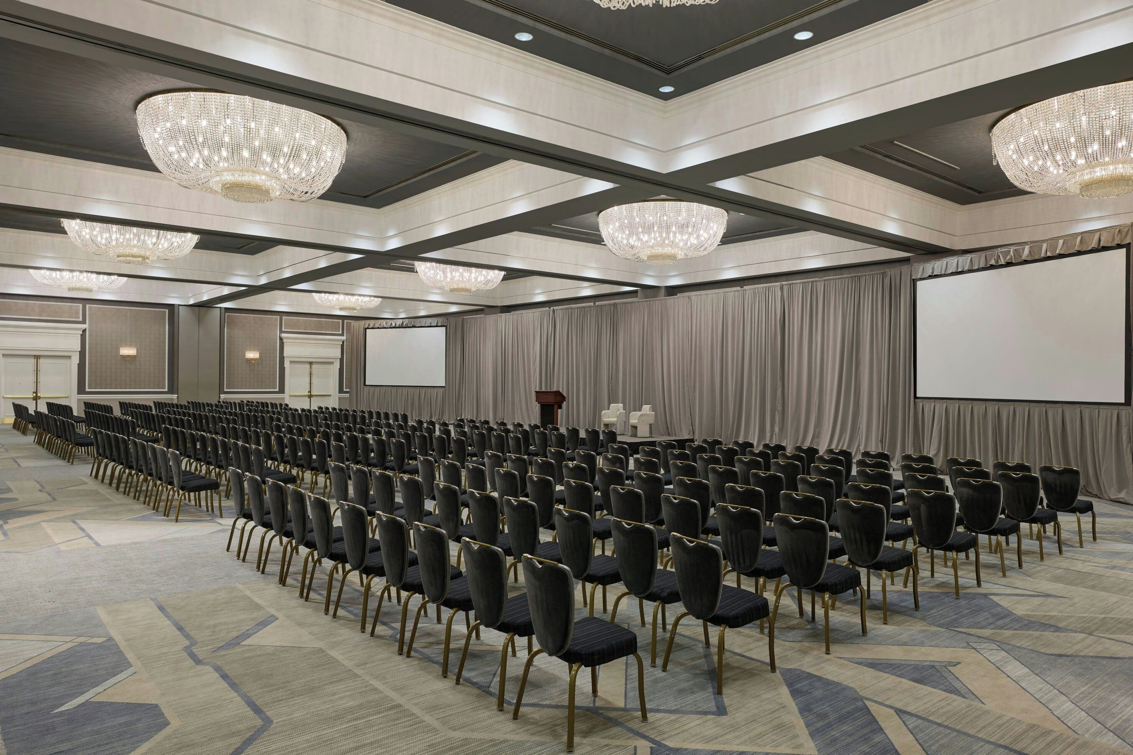 A conference room with rows of chairs, a large screen, and chandeliers.