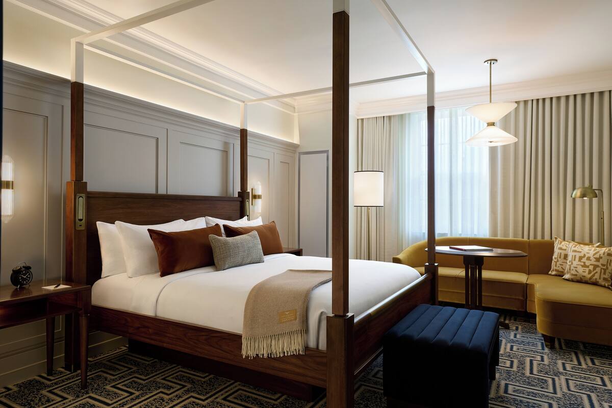 A four-poster bed with a wooden frame, a bedside table with a lamp, a sofa, and a small round table with a lamp.