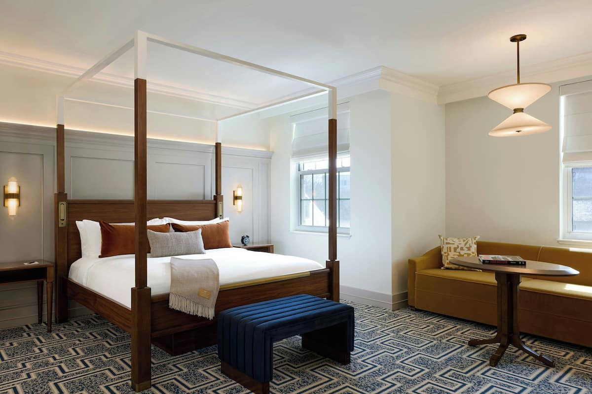 A four-poster bed with a wooden frame and white bedding, a small bedside table with a lamp, a bench, and a sofa with a patterned cushion.