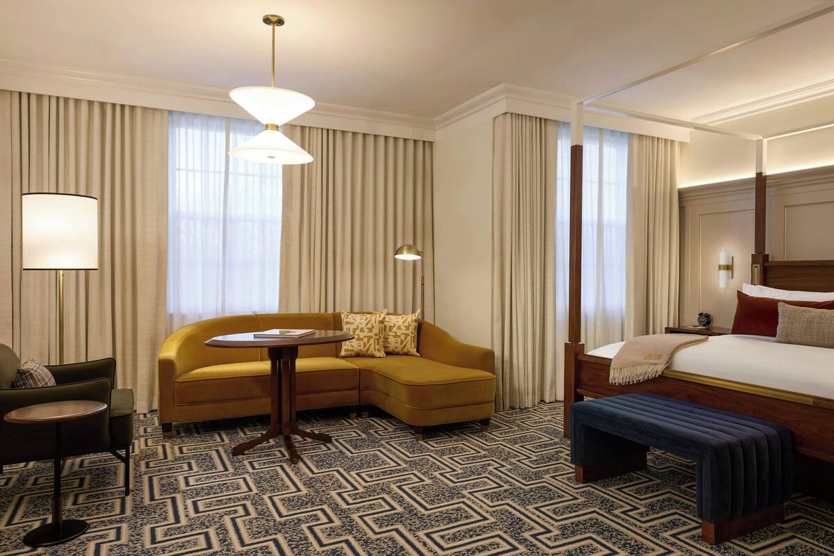A hotel room with a bed, a sofa, a small table, a bench, and a lamp.