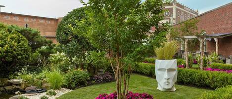 Ein angelegter Garten mit verschiedenen Pflanzen und Blumen, einem markanten Skulpturelement und einem Gebäude mit Backsteinfassade im Hintergrund.