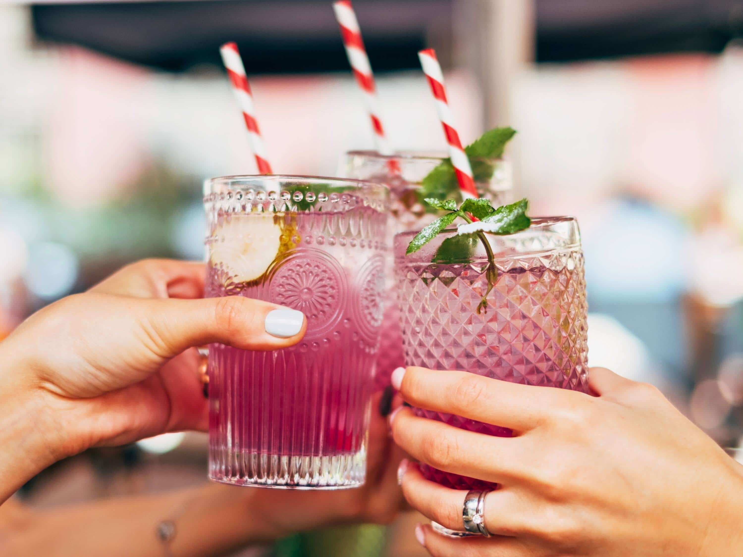 Two hands holding glasses of a pink beverage with straws and mint leaves.