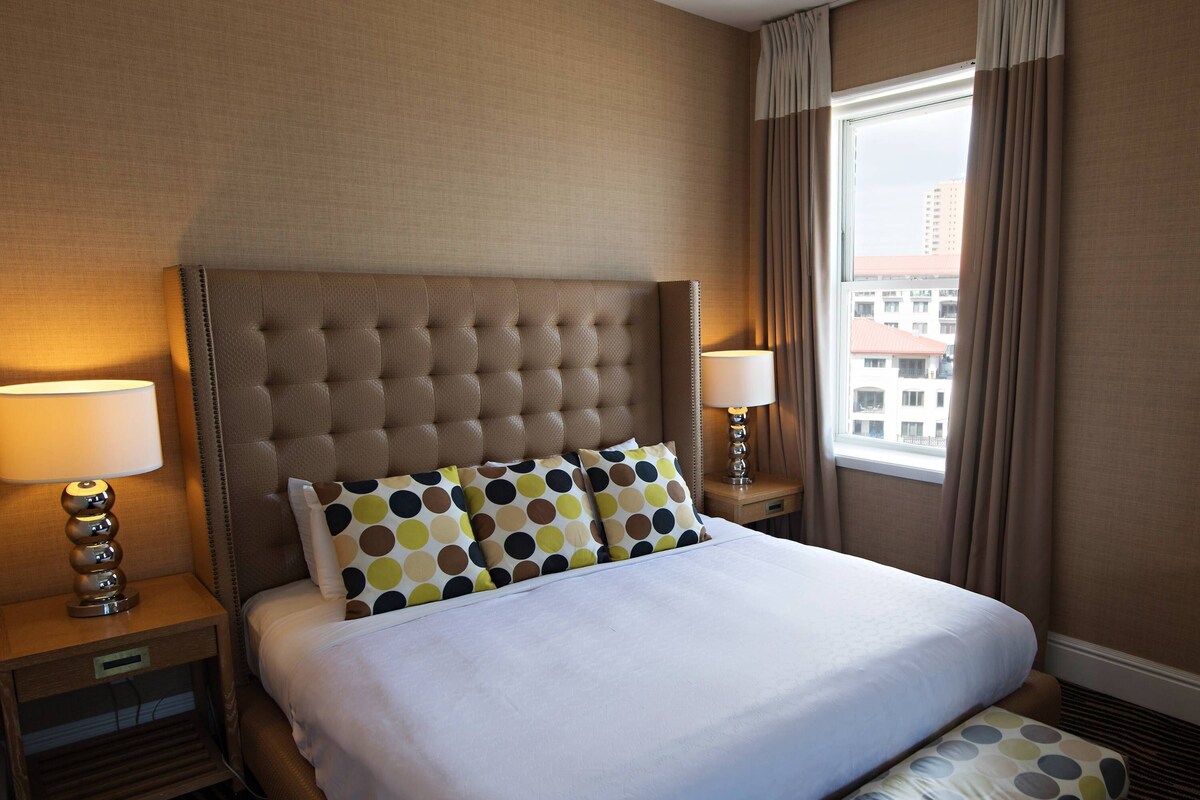 A hotel room with a large bed, two bedside lamps, a window with curtains, and a view of buildings outside.