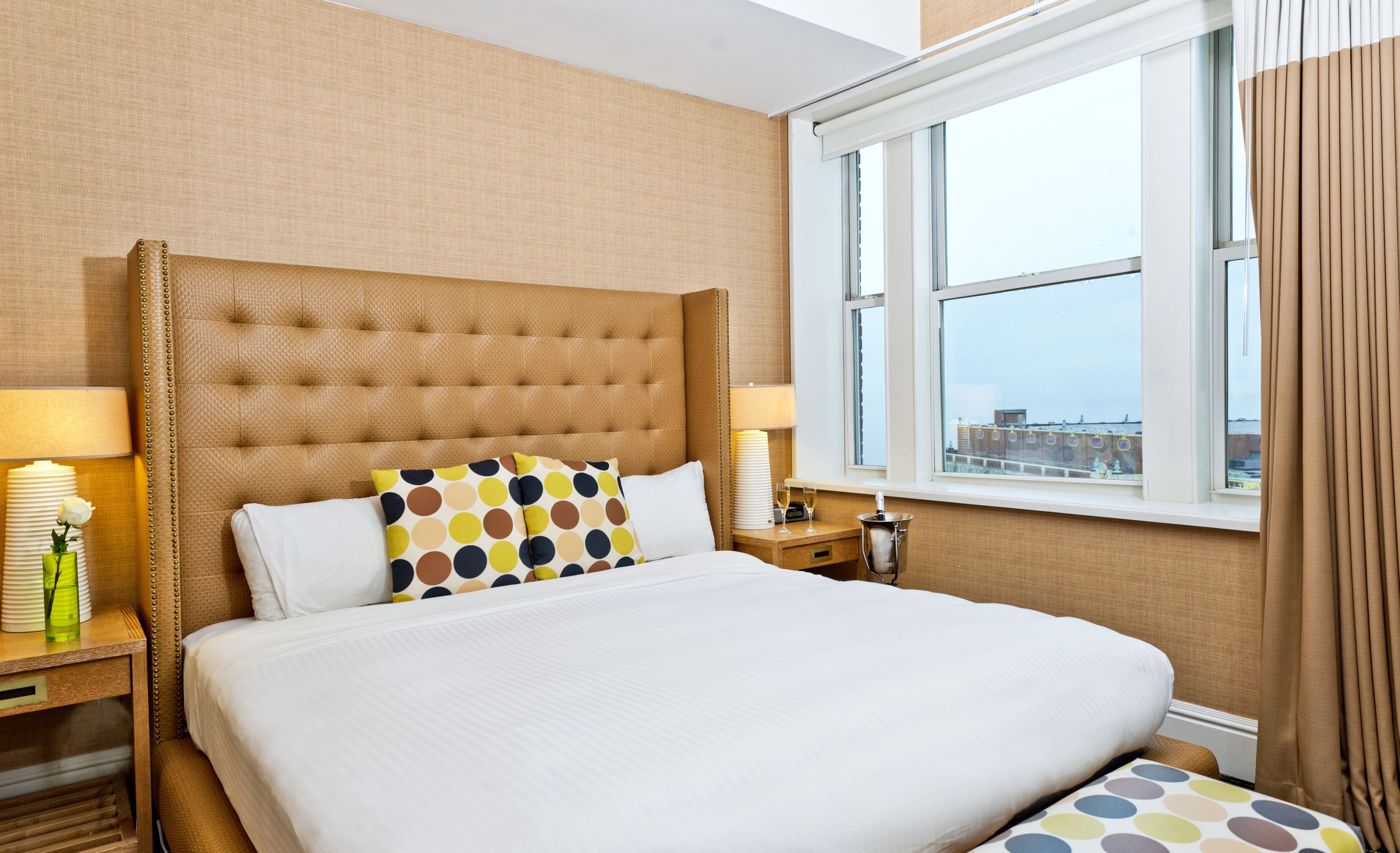A hotel room with a large bed, two bedside tables, a window with curtains, and a view of buildings.