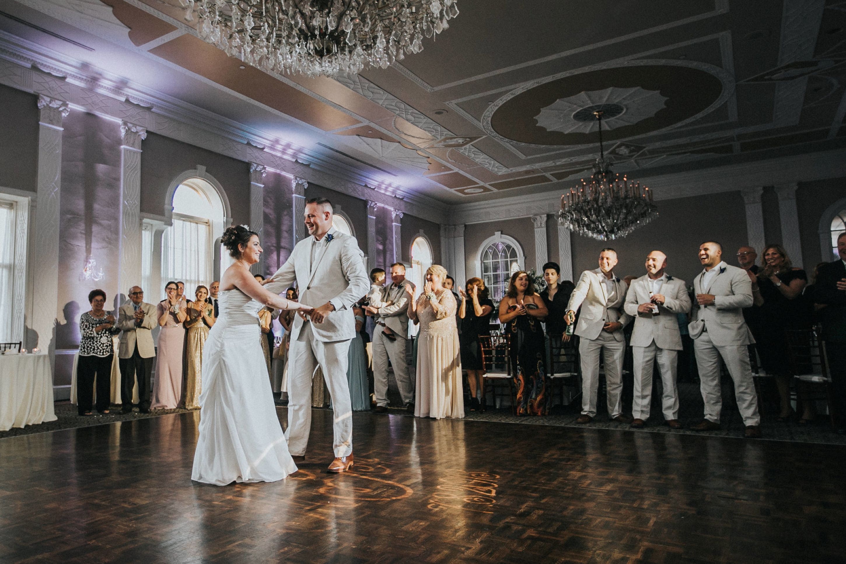 Una recepción de boda en un salón elegante con candelabro, suelo de madera y invitados de pie en semicírculo.