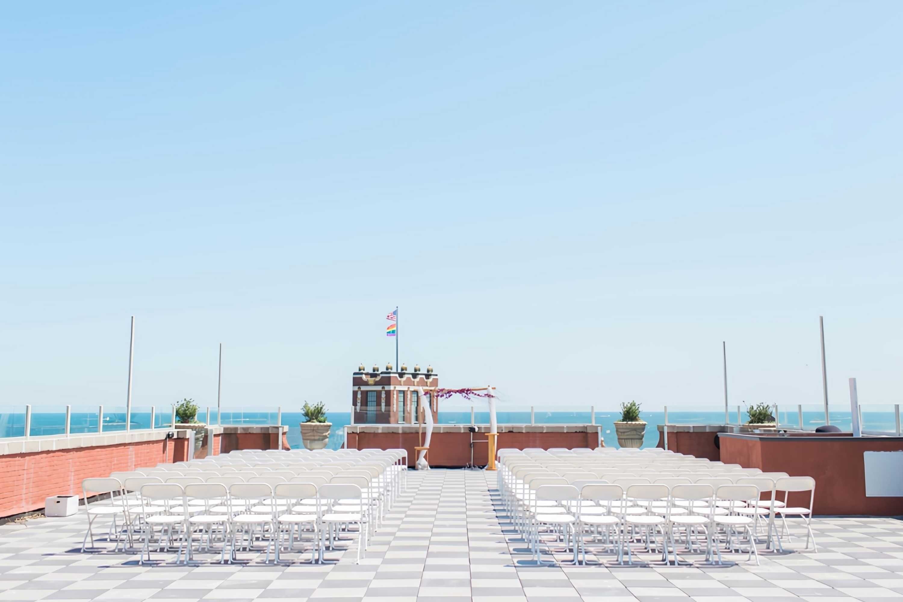 Un espace extérieur aménagé avec des chaises et des tables blanches, un drapeau et une vue sur la mer.