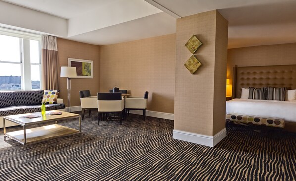 A hotel room with a sofa, a dining table, a coffee table, a bed, and a view of the city through large windows.