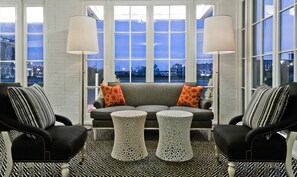 A modern living room with a grey sofa, two armchairs, and a patterned rug. Large windows offer a view of the cityscape.