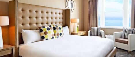 A hotel room with a large bed, a nightstand, a chair, a lamp, and a window with a view of the sea.