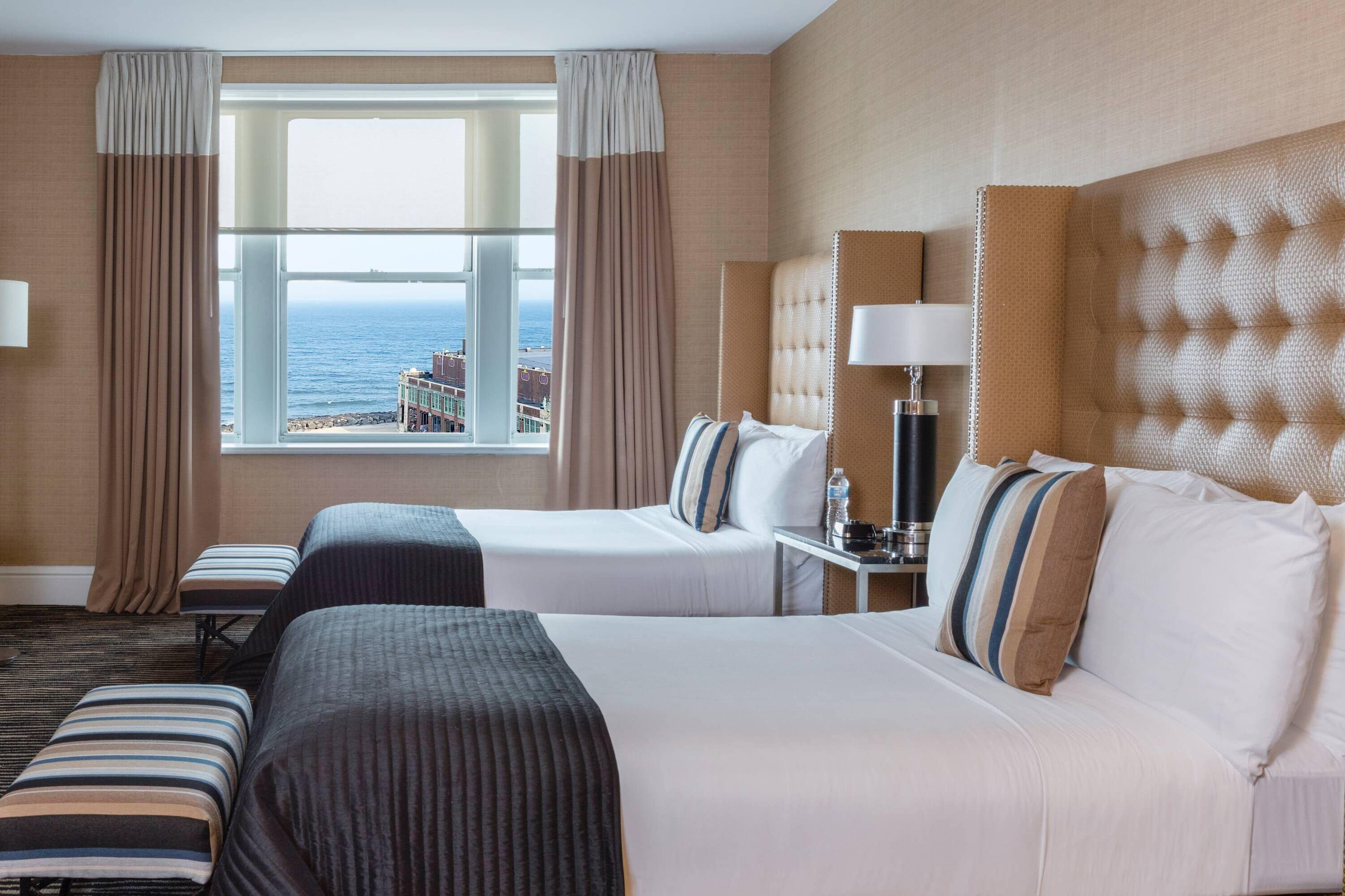 A hotel room with two beds, a view of the ocean, and a window with curtains.