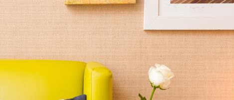 A yellow sofa with a polka dot pillow, a white rose in a green vase, and two abstract paintings on the wall.