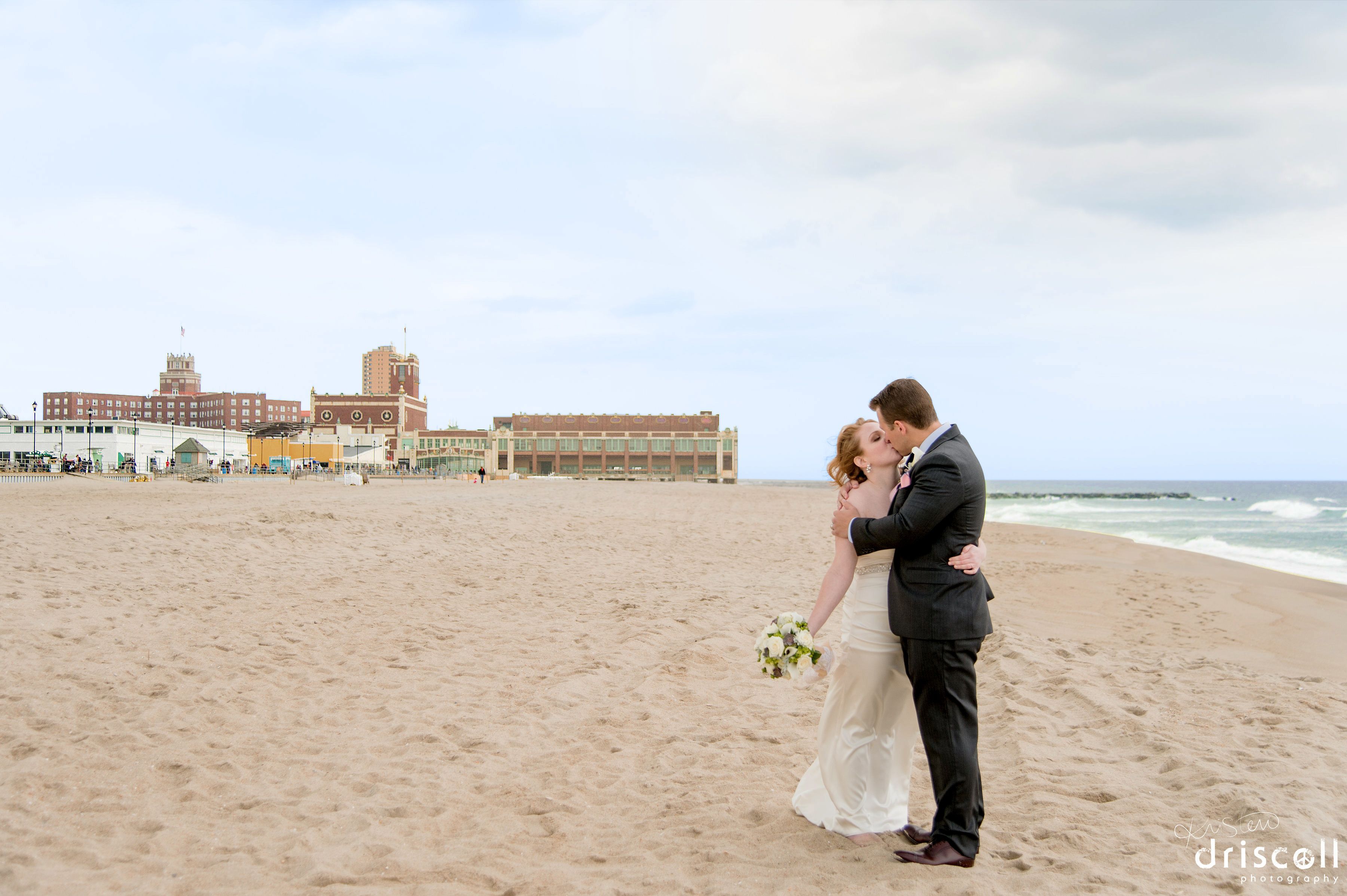 Escena de boda en la playa con una pareja abrazándose, un edificio con torre de reloj al fondo y el océano.