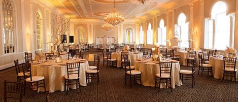 A banquet hall with round tables, chandeliers, and arched windows.