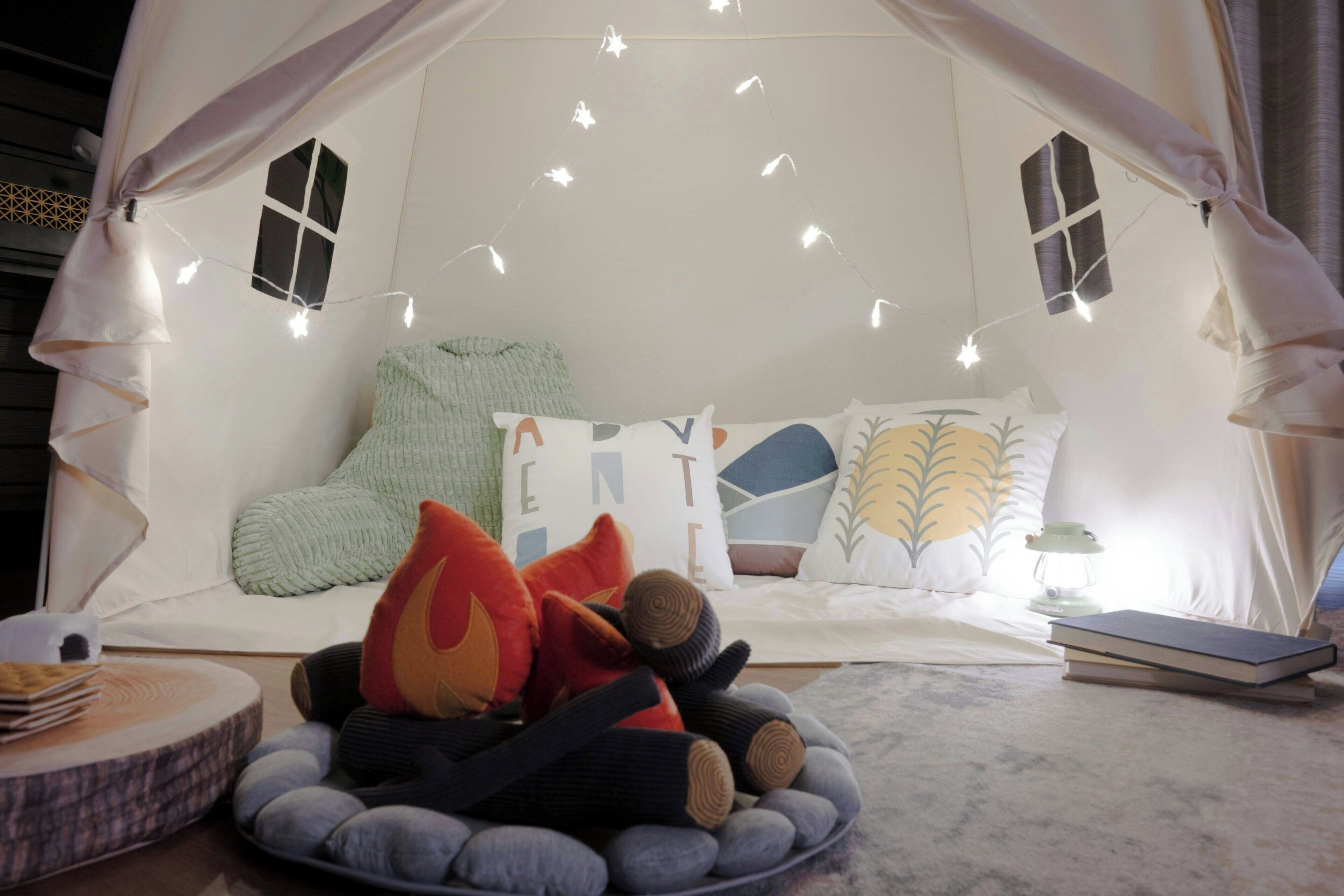 Ein gemütlicher, zeltartiger Raum mit Lichterketten, einem Bett mit Kissen, einem kleinen Tisch mit Büchern und einer Lampe.