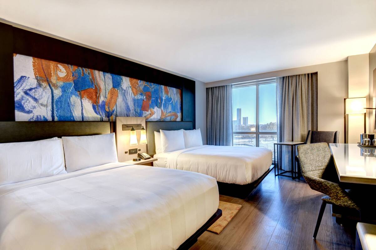 A hotel room with two beds, a dining table, a chair, and a large abstract painting on the wall.