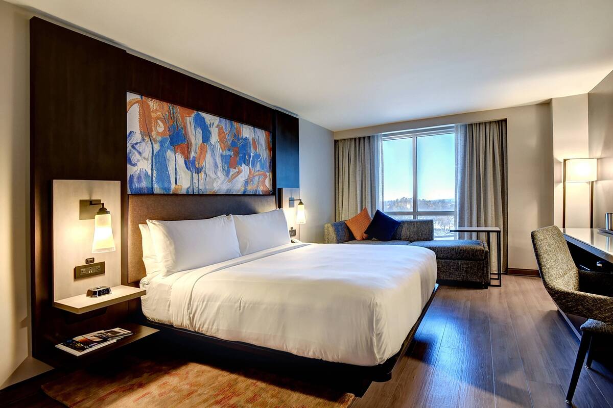A hotel room with a large bed, a desk, a chair, a nightstand, a lamp, and a painting on the wall.