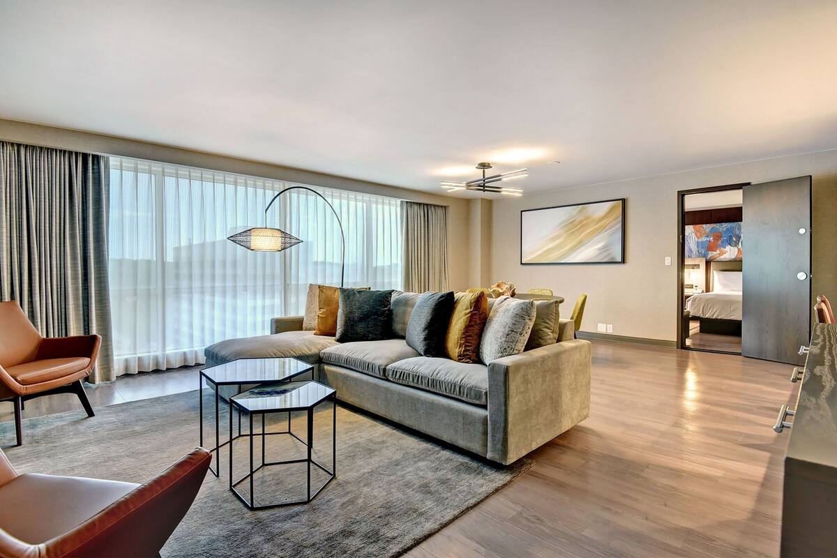 A modern living room with a sofa, armchairs, and a coffee table. There is a large window with curtains, a ceiling fan, and a painting on the wall.