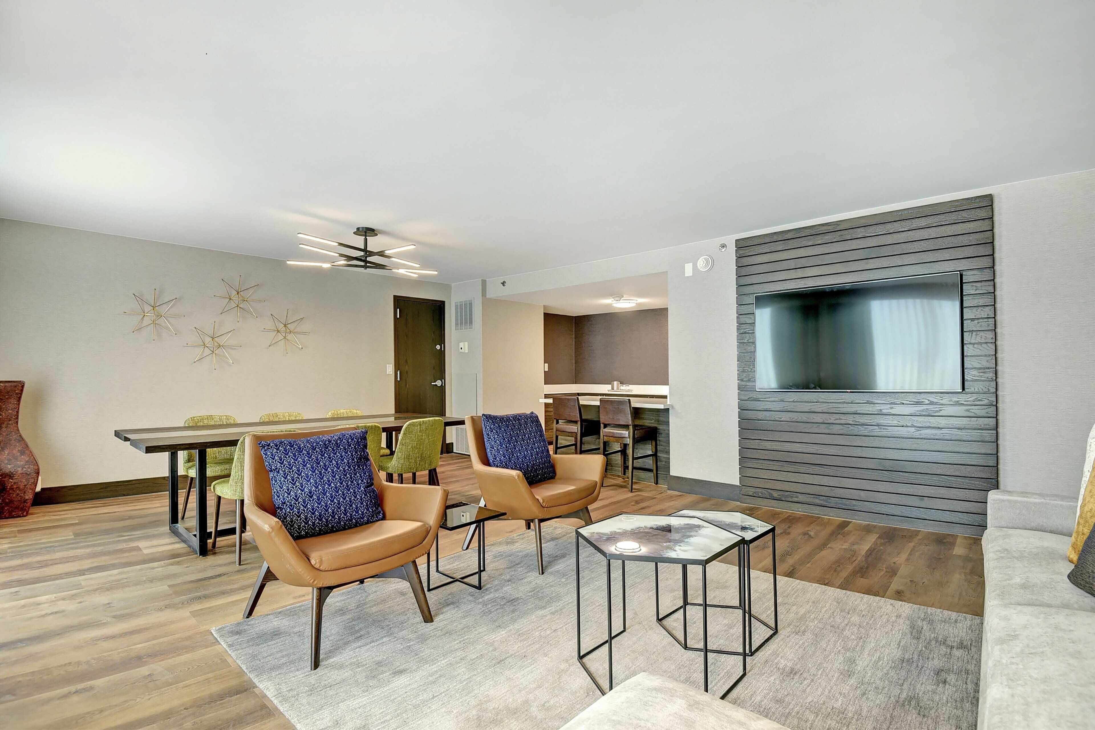 A modern living room with a dining area, featuring leather chairs, a glass coffee table, and a large flat-screen TV mounted on a wooden panel wall.