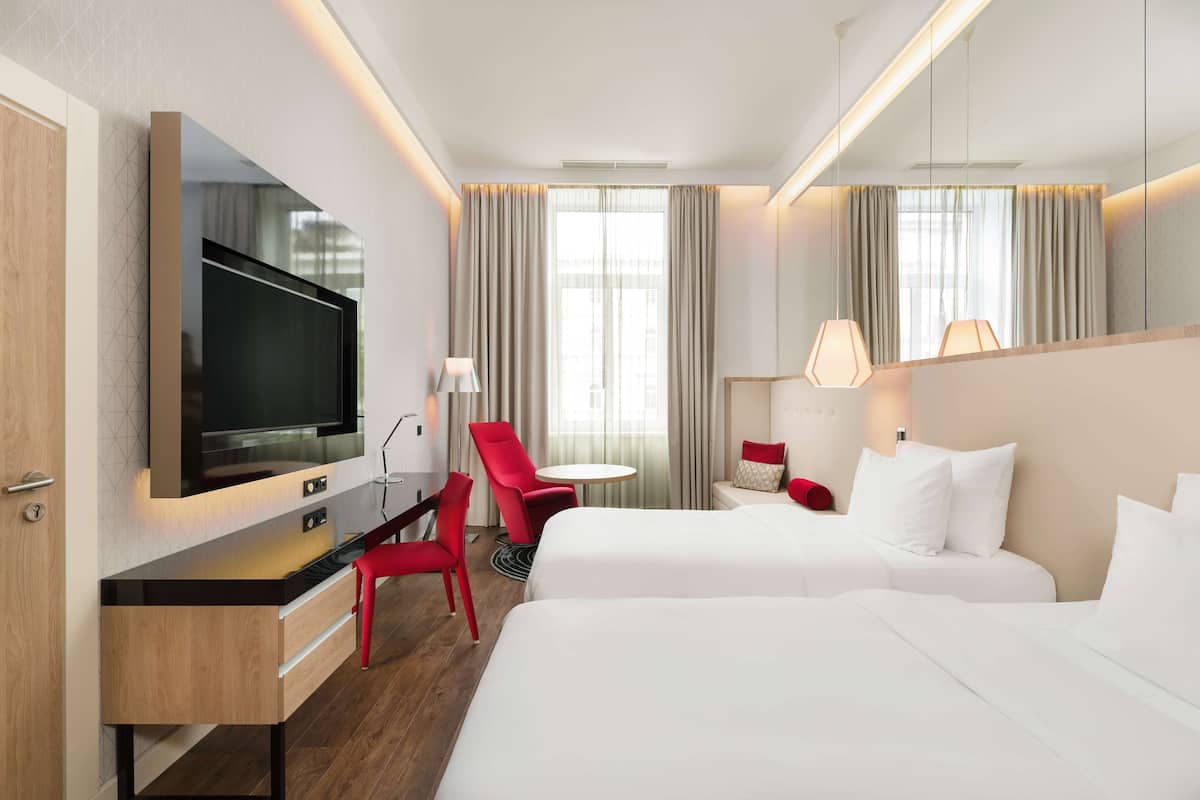 Habitación de hotel con televisor de pantalla plana, escritorio con silla, dos camas, una mesita pequeña y una lámpara.