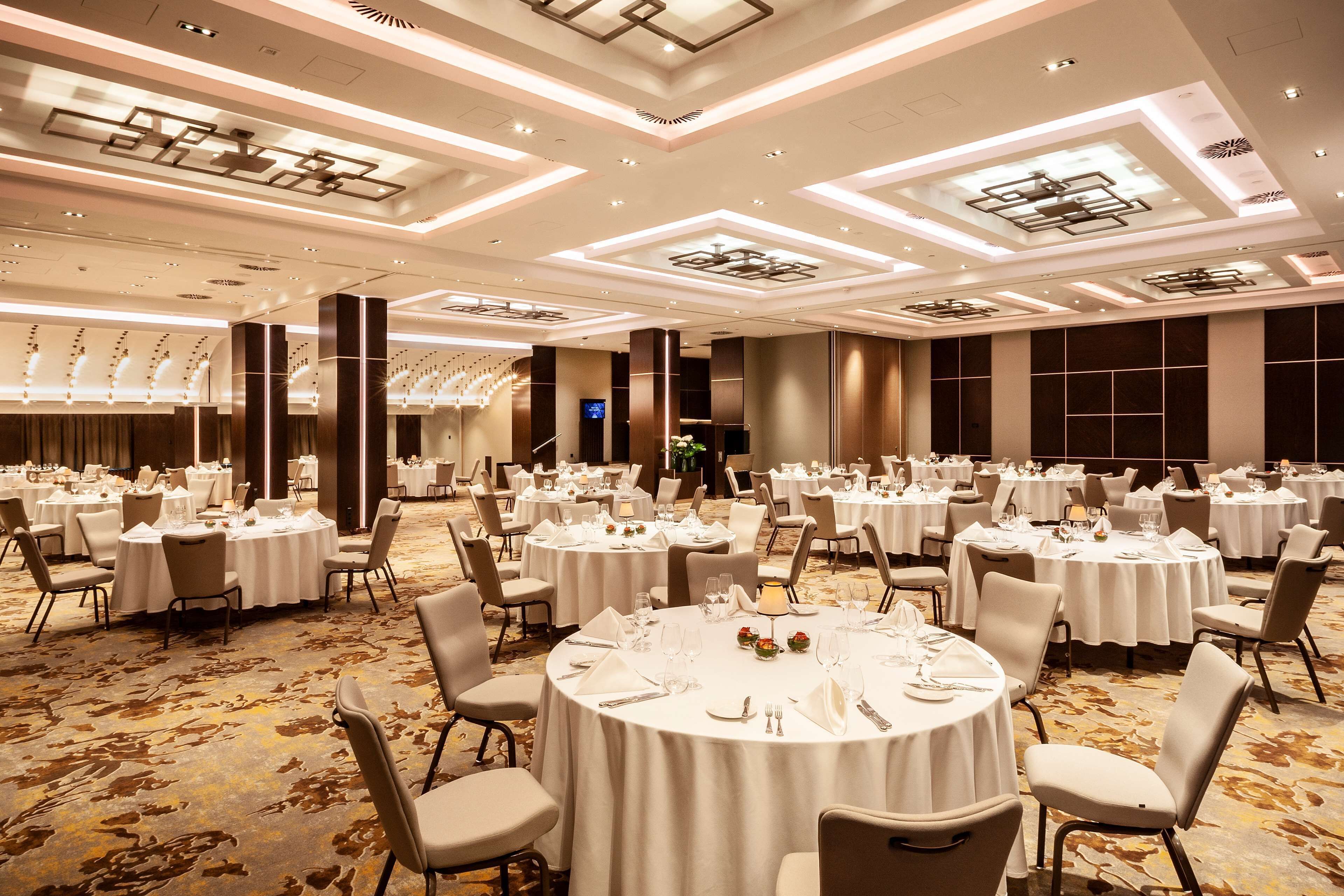 Une grande salle de banquet avec des tables rondes dressées pour le repas, dotée d’un plafond au design moderne et d’un sol recouvert de moquette.