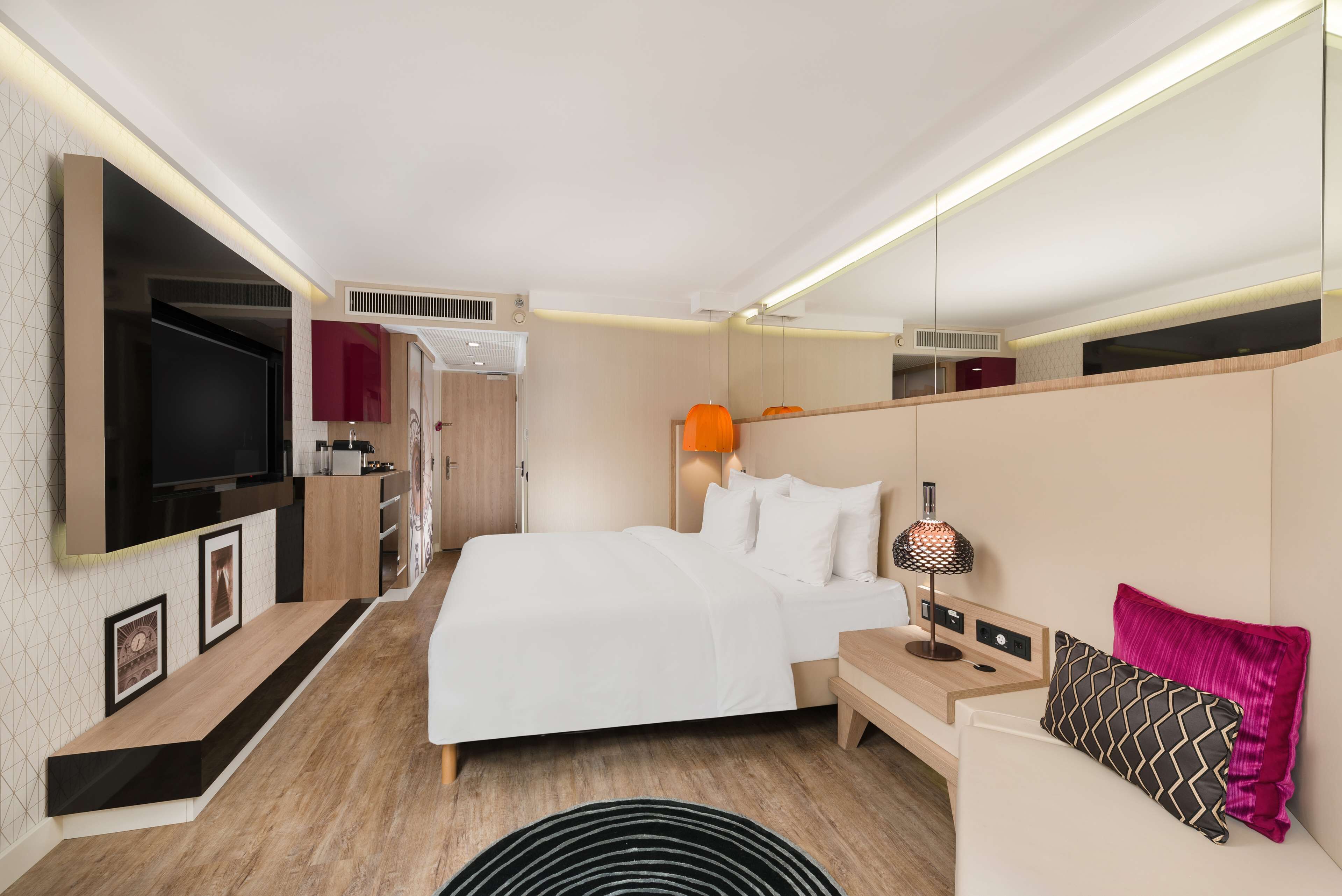 Una habitación de hotel moderna con una cama grande, un televisor montado en la pared, una mesita de noche con lámpara y una zona de estar con sofá.