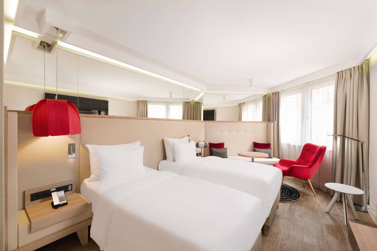 Una habitación de hotel con dos camas, una silla roja, una mesita y una lámpara.