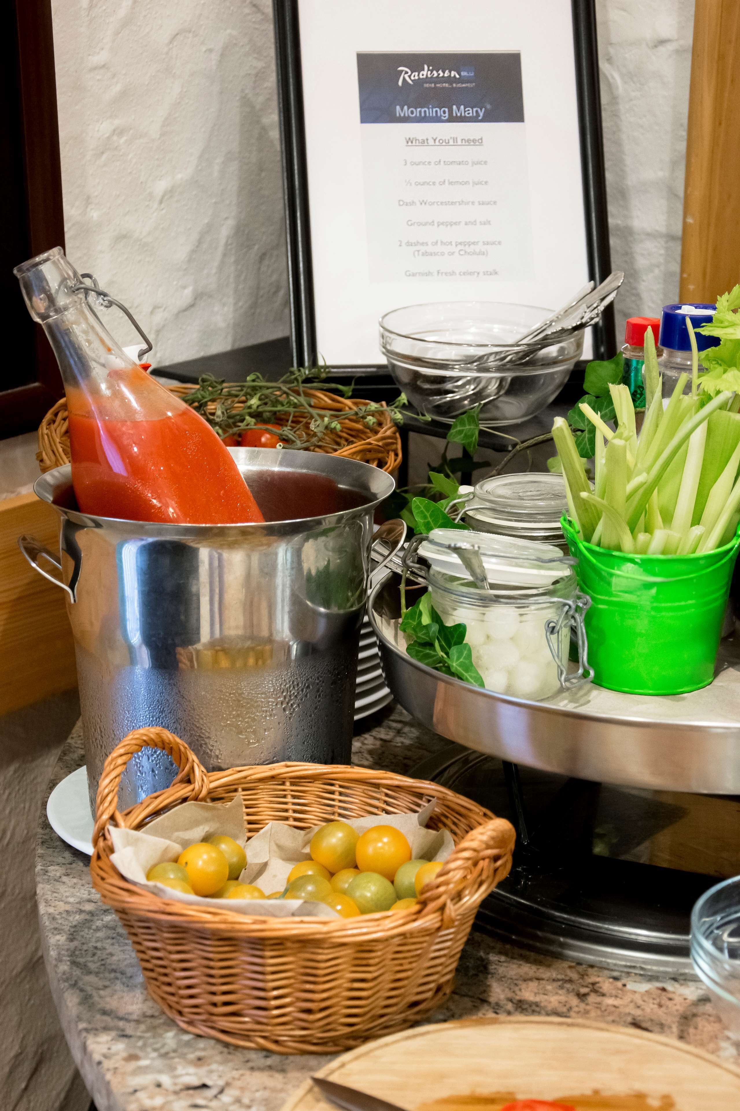 A kitchen counter with various ingredients and utensils, including a basket of yellow fruits, a glass container with celery, and a metal pot with a tomato-based sauce.