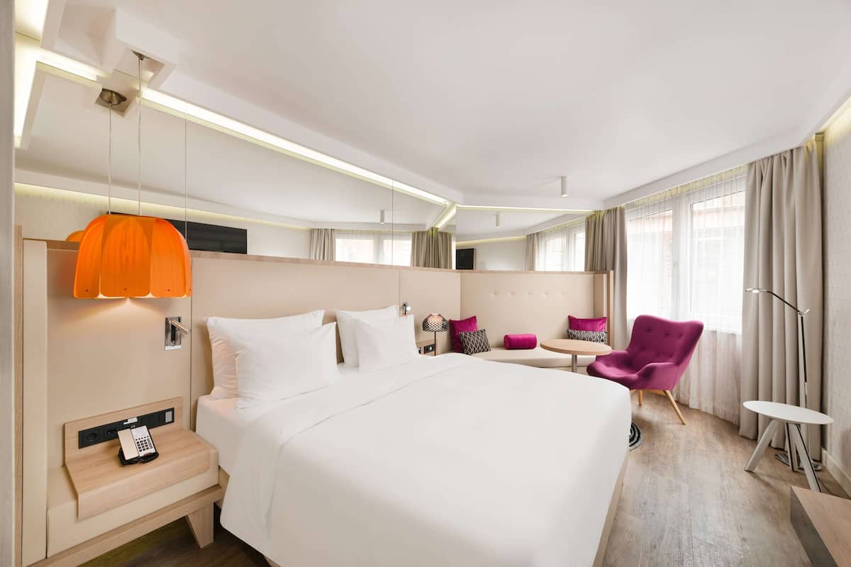 Una habitación de hotel moderna con una cama grande, mesita de noche con teléfono, una silla, una pequeña mesa y una lámpara.
