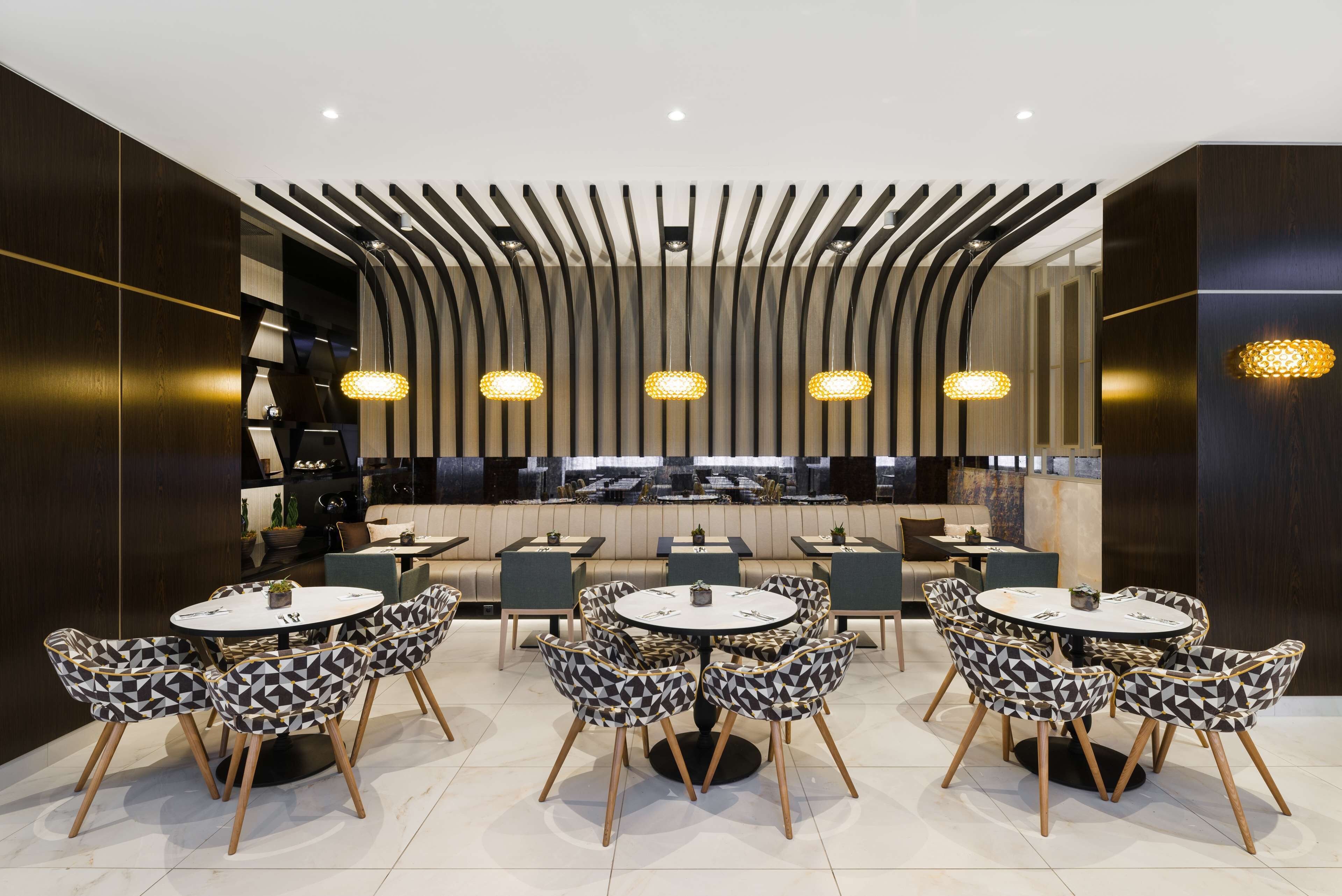 A modern restaurant with round tables and patterned chairs, featuring a unique ceiling design and pendant lighting.