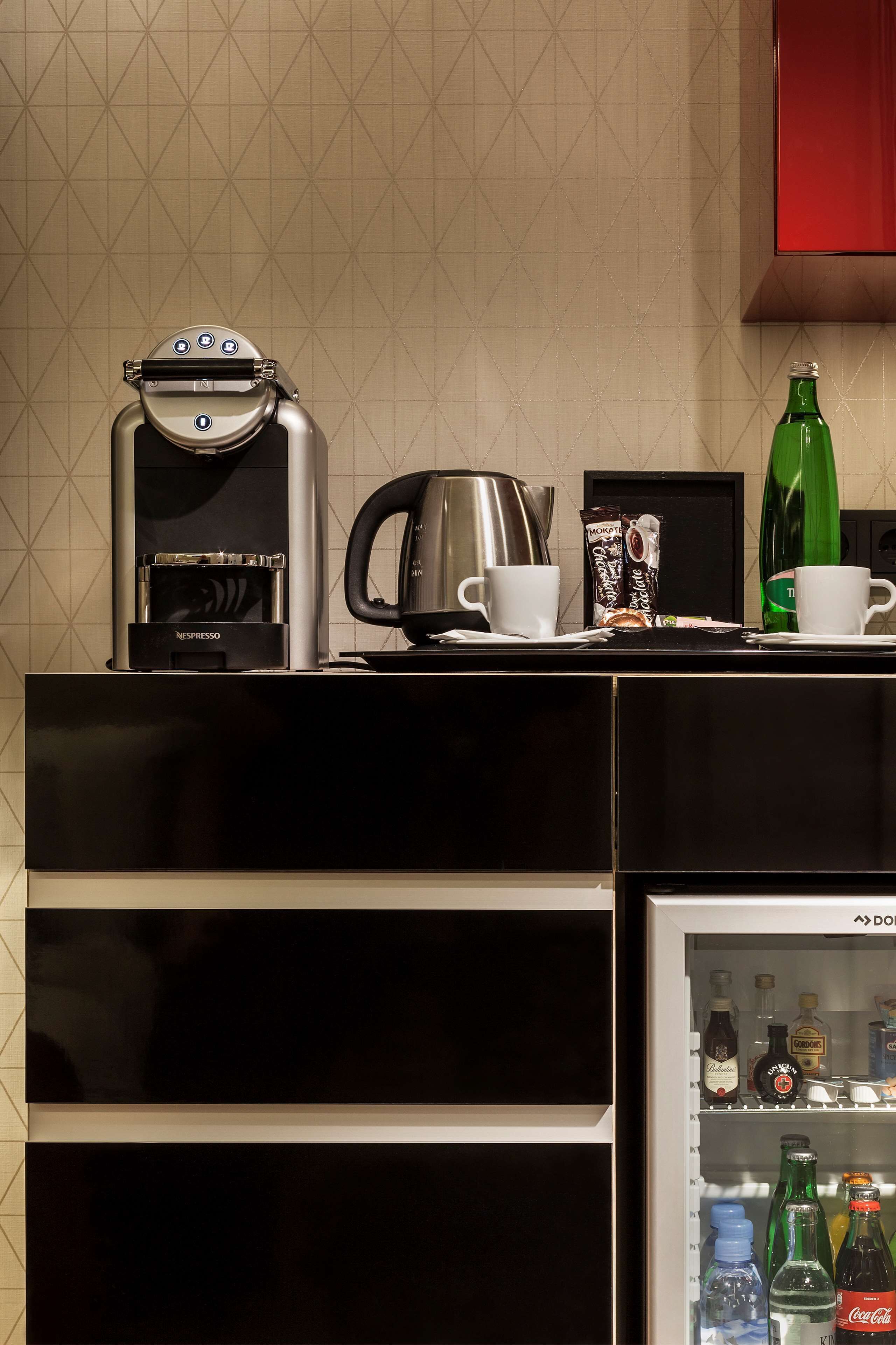 Un comptoir de cuisine avec une machine à café, une bouilloire, deux tasses, une bouteille verte et un réfrigérateur rempli de boissons.