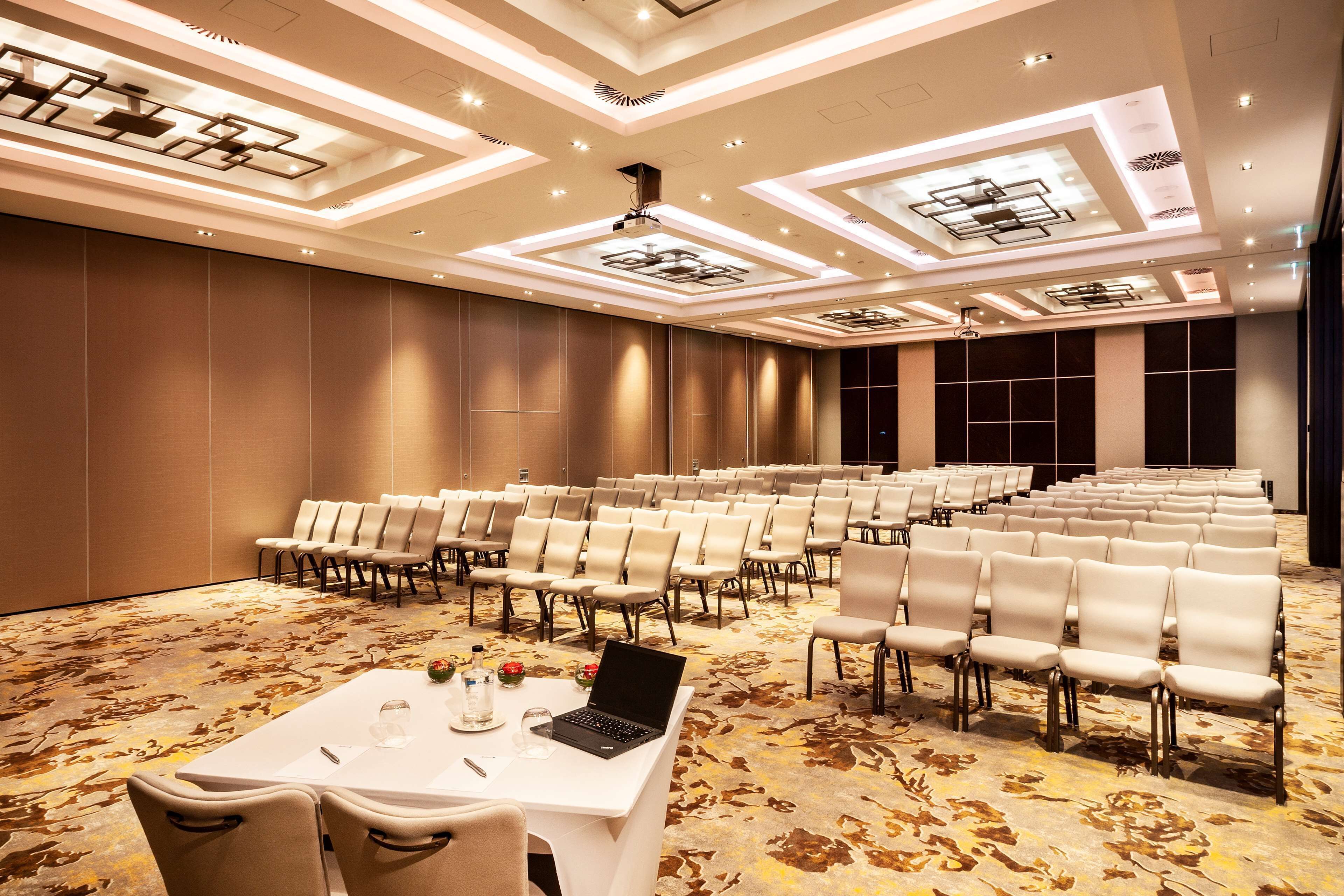 A conference room with rows of chairs, a table with a laptop, and a patterned carpet.
