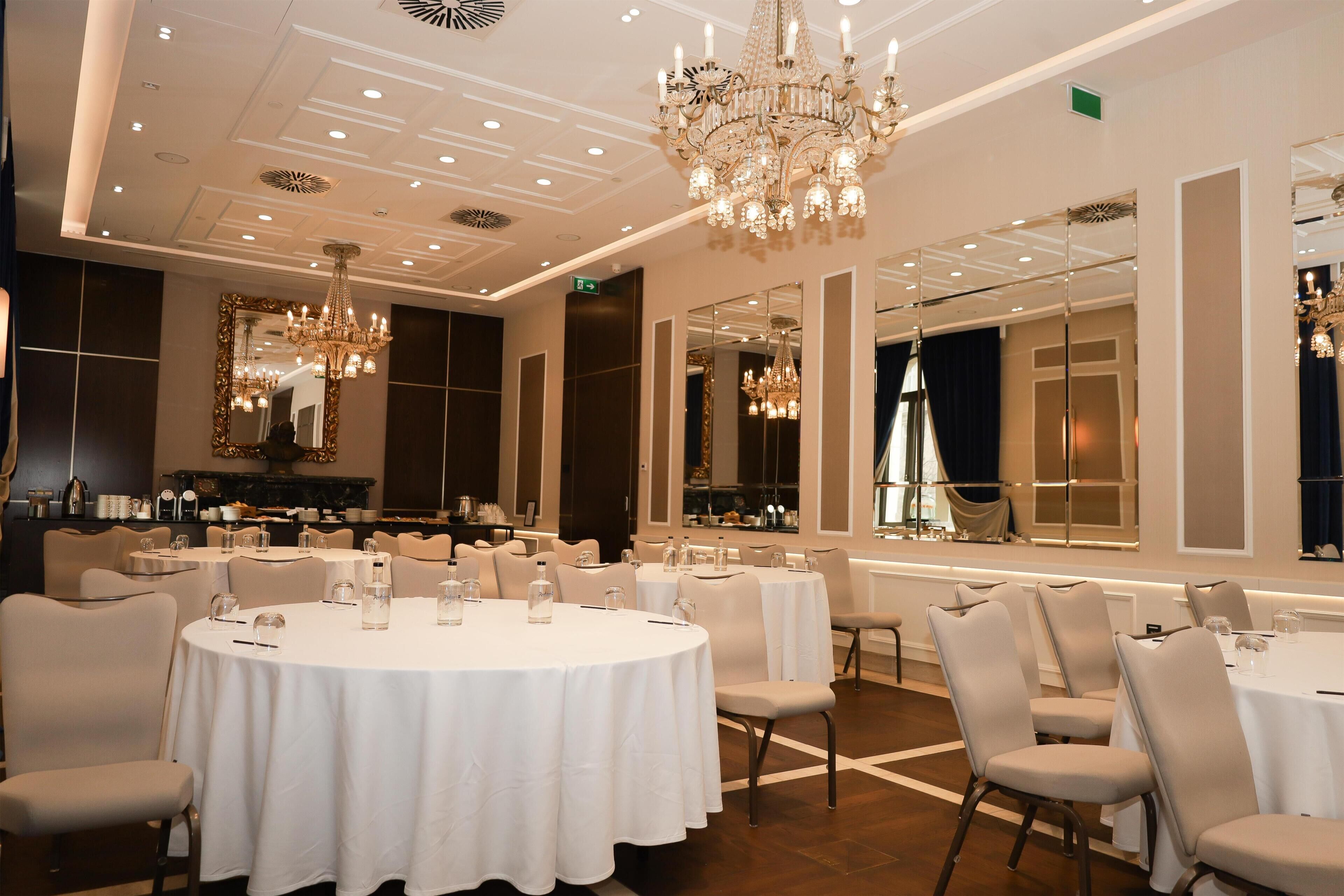 A formal dining room with round tables, chairs, chandeliers, and large mirrors.
